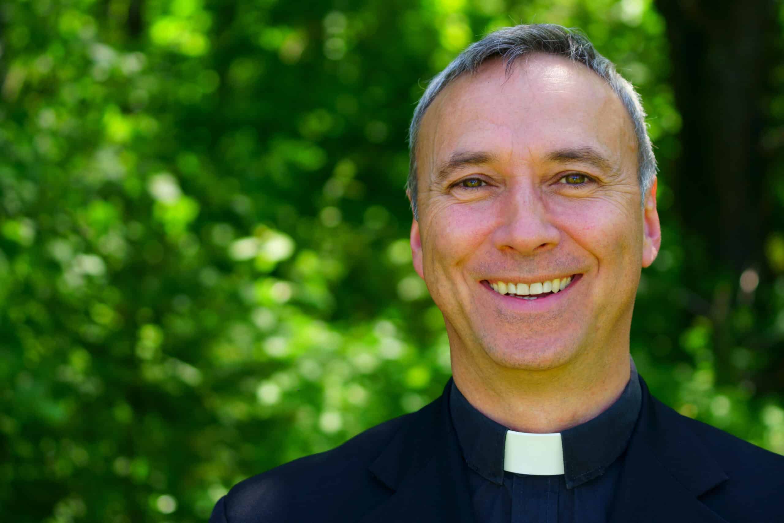 A good looking catholic priest is standing in the forest, smiling at us.